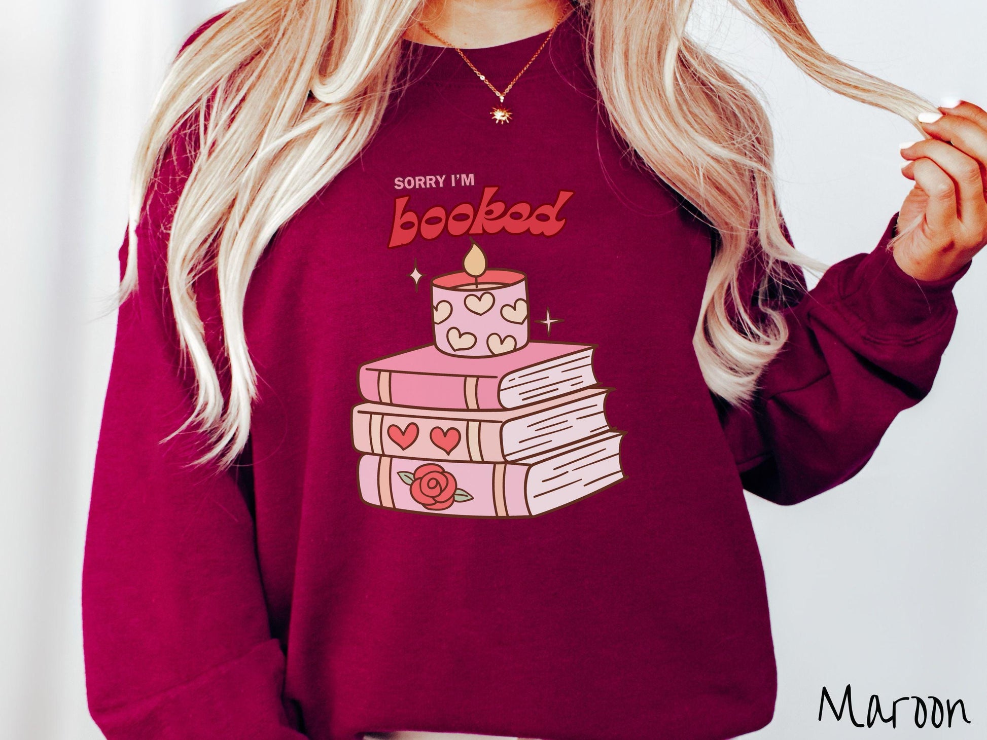 A vintage Gildan sweatshirt featuring the playful phrase “Sorry I’m Booked” in pink and red font, completed with a stack of three pink and purple books adorned with red roses and hearts on their spines, topped with a lit candle.