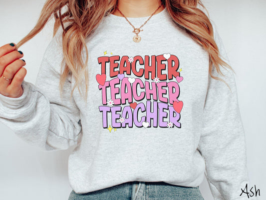 A vibrant vintage Gildan sweatshirt featuring the word “Teacher” written vertically three times in bold red, pink, and purple font, with colorful hearts, flowers, and twinkling stars scattered throughout for a whimsical and eye-catching look.