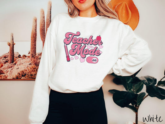 A vintage Gildan sweathirt featuring the phrase “Teacher Mode” in playful pink font, an “On” button toggled to the “On” position, and with charming details like a red pencil, a red apple, and pink hearts sprinkled throughout.