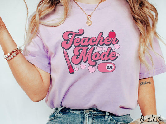 A vintage Comfort Colors t-shirt featuring the phrase “Teacher Mode” in playful pink font, an “On” button toggled to the “On” position, and with charming details like a red pencil, a red apple, and pink hearts sprinkled throughout.