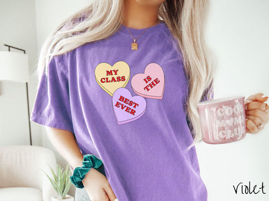 A charming vintage-style Valentine’s Day Comfort Colors t-shirt featuring three vibrant conversation hearts in yellow, red, and purple, this design displays the text “My class is the best ever” spread across the hearts.