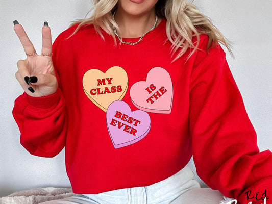 A charming vintage-style Valentine’s Day Gildan sweatshirt featuring three vibrant conversation hearts in yellow, red, and purple, this design displays the text “My class is the best ever” spread across the hearts.