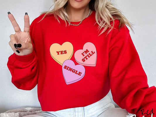 Make a bold statement this Valentine’s Day with this vintage Gildan sweatshirt featuring three vibrant conversation hearts in yellow, red, and purple, the design showcasing the cheeky phrase “Yes, I’m Still Single” spread across the hearts.