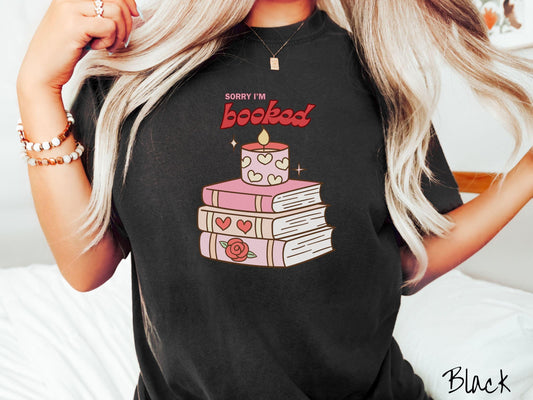 A vintage Comfort Colors t-shirt featuring the playful phrase “Sorry I’m Booked” in pink and red font, completed with a stack of three pink and purple books adorned with red roses and hearts on their spines, topped with a lit candle.