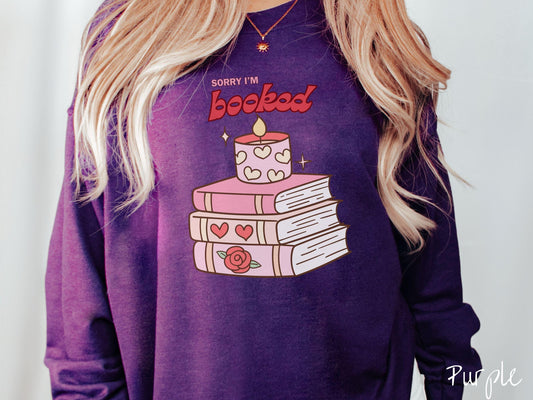 A vintage Gildan sweatshirt featuring the playful phrase “Sorry I’m Booked” in pink and red font, completed with a stack of three pink and purple books adorned with red roses and hearts on their spines, topped with a lit candle.
