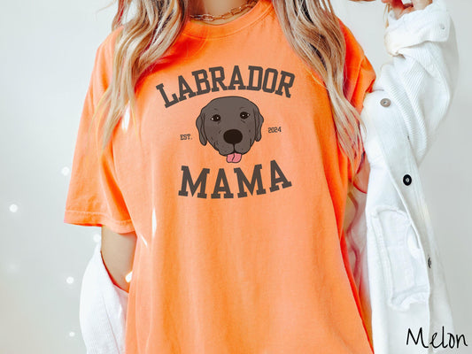 A vintage Comfort Colors t-shirt featuring bold block lettering that reads &#39;Labrador Mama.&#39; At the center of the design, a playful black Labrador with its tongue out steals the spotlight, framed by the stylish text &#39;EST. 2024.&#39;