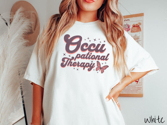 A vintage, white colored Comfort Colors t-shirt featuring the words Occupational Therapy across the front, this unique design showcases a stunning pink and dark purple butterfly surrounded by delicate twinkling stars.