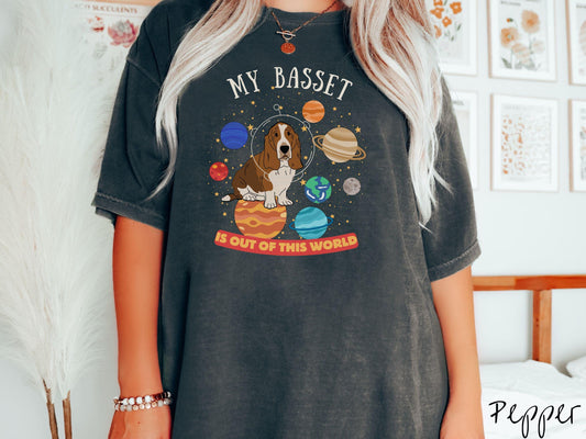 A vintage Comfort Colors t-shirt featuring the playful text &#39;My Basset is Out of This World&#39;. The design showcases a basset hound sitting proudly atop Jupiter, surrounded by a vibrant solar system filled with colorful planets and twinkling stars.