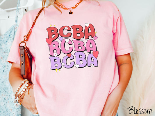 A vibrant Comfort Colors T-shirt with the text BCBA written three times vertically, accompanied by tiny hearts, stars, and flowers. Perfect for Board Certified Behavior Analysts, this tee is a great Valentine&#39;s Day gift to show appreciation and love.