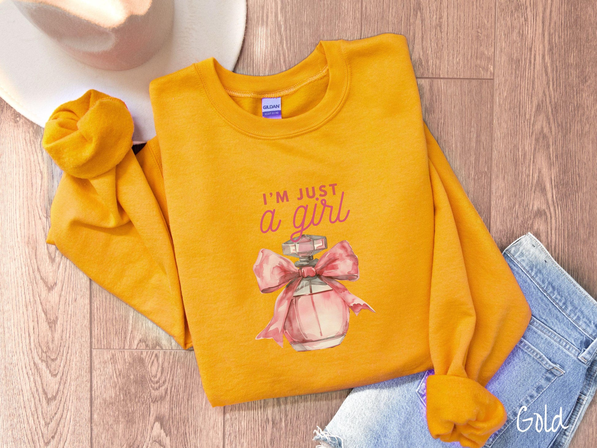 A cozy Gildan sweatshirt featuring the phrase &quot;I&#39;m Just a Girl&quot; in pink font above a bottle of pink perfume tied with a pink bow. Perfect for those colder months, this sweatshirt provides warmth and comfort with its classic fit and crew neckline.