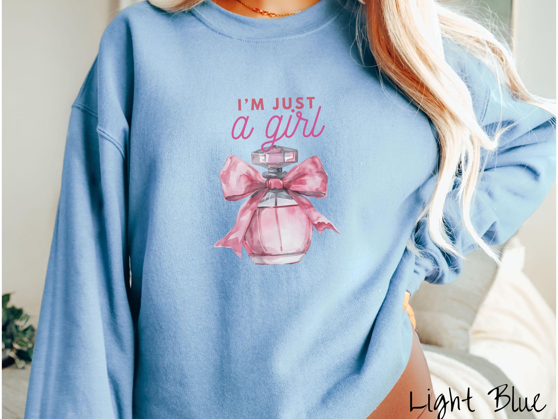 A cozy Gildan sweatshirt featuring the phrase &quot;I&#39;m Just a Girl&quot; in pink font above a bottle of pink perfume tied with a pink bow. Perfect for those colder months, this sweatshirt provides warmth and comfort with its classic fit and crew neckline.