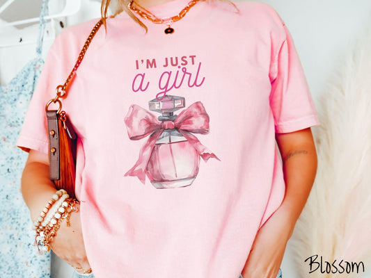 A stylish Comfort Colors T-shirt featuring the phrase &quot;I&#39;m Just a Girl&quot; in pink font above a bottle of pink perfume tied with a pink bow. This tee exudes a feminine and trendy vibe, perfect for those who appreciate unique fashion statements.