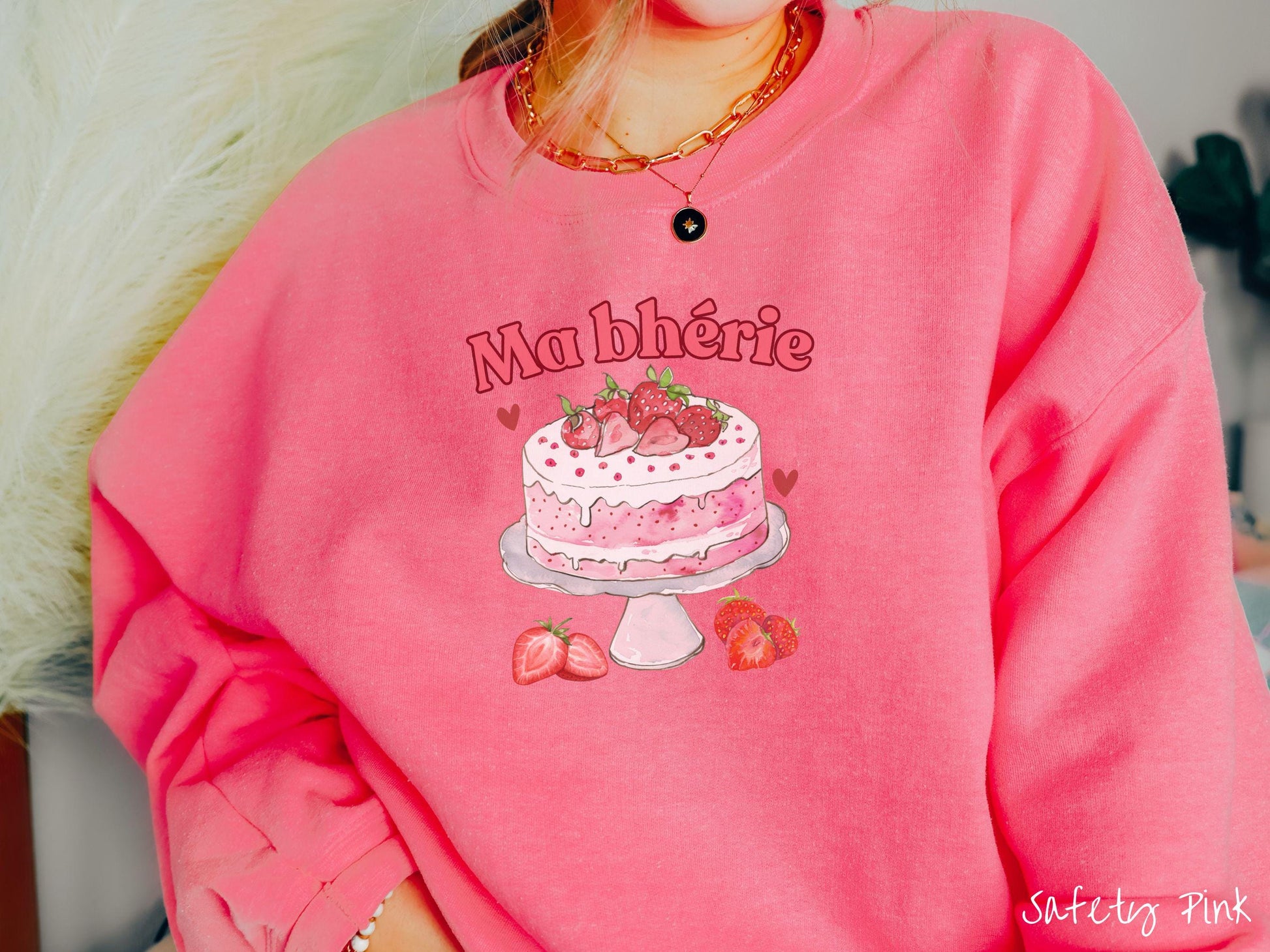 A Gildan sweatshirt featuring the phrase &quot;Ma Bherie&quot; is printed in bold red font above a charming illustration of a red-and-white strawberry cake displayed on a plate, topped with fresh strawberries and surrounded by even more berries.