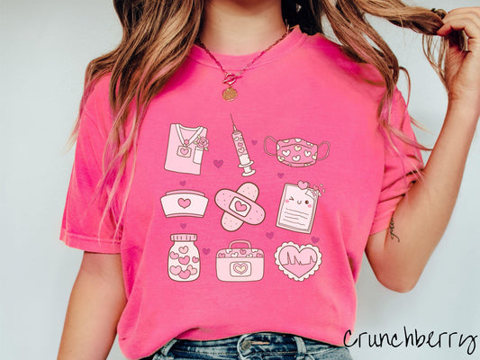 A Comfort Colors T-shirt featuring a 3x3 grid of nursing-themed illustrations adorned with heart designs like nursing scrubs, syringes, a face mask, a nurse cap, bandages, medical records, vitamins, a medical bag, and a heart-shaped EKG graph.