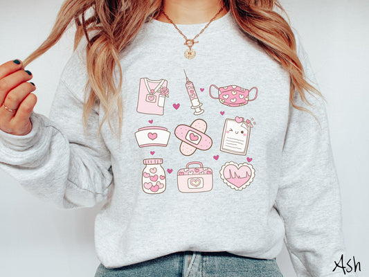 A Gildan sweatshirt featuring a 3x3 grid of nursing-themed illustrations adorned with heart designs like nursing scrubs, syringes, a face mask, a nurse cap, bandages, medical records, vitamins, a medical bag, and a heart-shaped EKG graph.