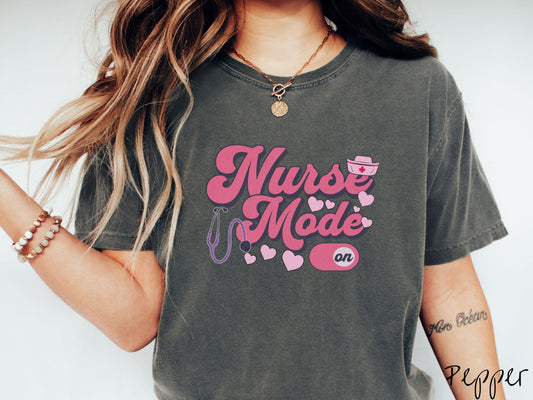 A stylish Comfort Colors t-shirt design features the text &quot;Nurse Mode&quot; in bold pink font with a pink nurse&#39;s cap placed over the letter E, and a stethoscope positioned beneath the text, alongside an &quot;on&quot; button widget set to the on position.