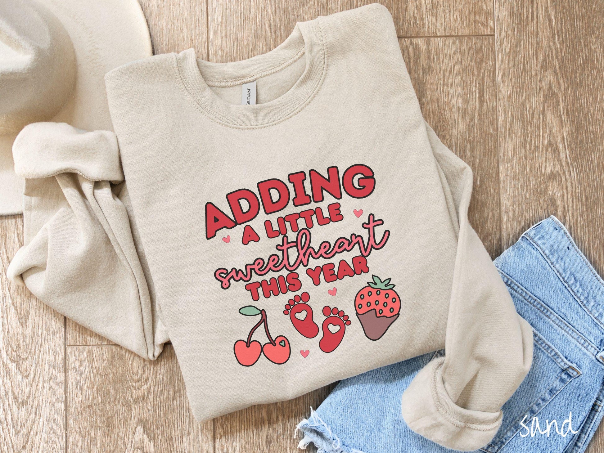 Share your exciting news with this vintage Gildan sweatshirt featuring the phrase “Adding a little sweetheart this year” in bold red font, paired with two red baby footprints framed by two red cherries and a chocolate-covered strawberry.