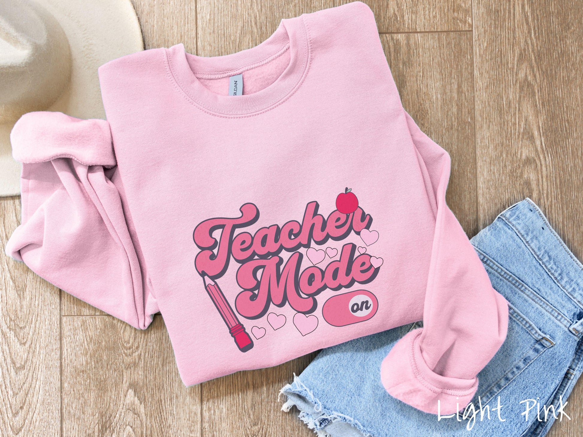 A vintage Gildan sweathirt featuring the phrase “Teacher Mode” in playful pink font, an “On” button toggled to the “On” position, and with charming details like a red pencil, a red apple, and pink hearts sprinkled throughout.