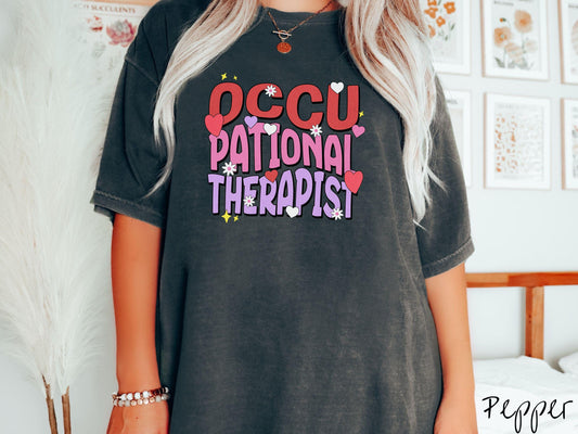 A stylish Comfort Colors t-shirt featuring the words &quot;Occupational Therapist&quot; arranged vertically in bold red, pink, and purple font, this design is enhanced with colorful hearts, flowers, and stars for a playful, uplifting touch.