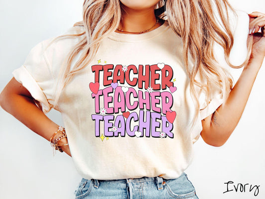 A vibrant vintage Comfort Colors t-shirt featuring the word “Teacher” written vertically three times in bold red, pink, and purple font, with colorful hearts, flowers, and twinkling stars scattered throughout for a whimsical and eye-catching look.