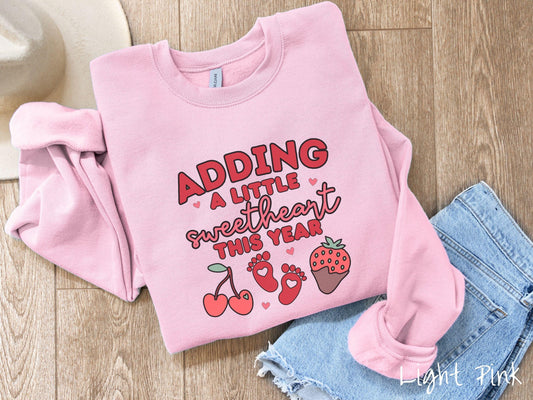 Share your exciting news with this vintage Gildan sweatshirt featuring the phrase “Adding a little sweetheart this year” in bold red font, paired with two red baby footprints framed by two red cherries and a chocolate-covered strawberry.