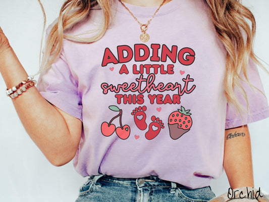 Share your exciting news with this vintage Comfort Colors t-shirt featuring the phrase “Adding a little sweetheart this year” in bold red font, paired with two red baby footprints framed by two red cherries and a chocolate-covered strawberry.