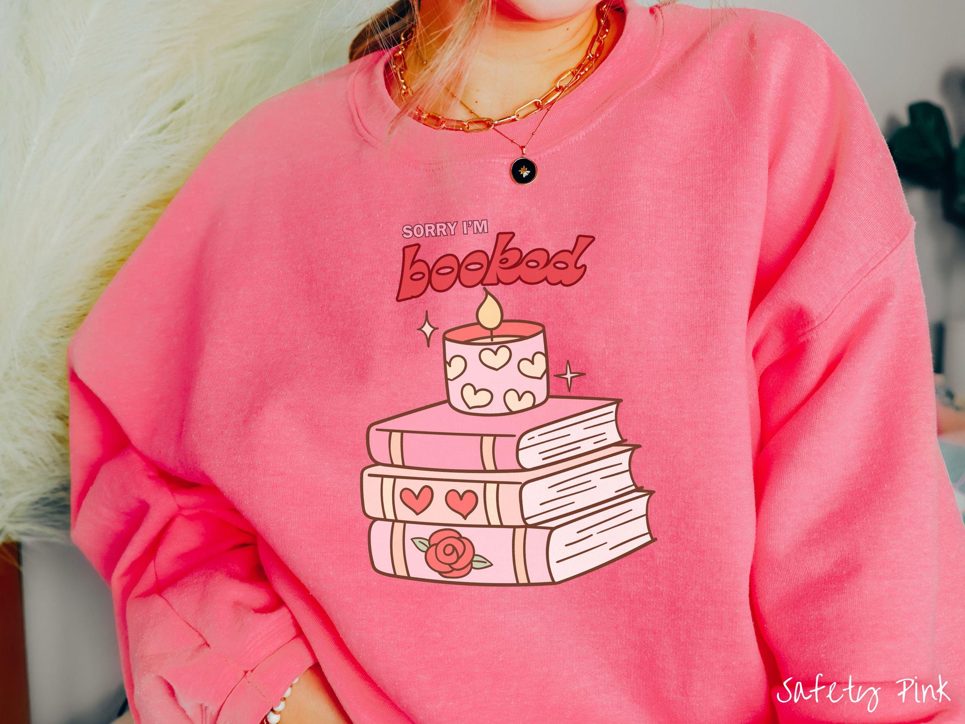 A vintage Gildan sweatshirt featuring the playful phrase “Sorry I’m Booked” in pink and red font, completed with a stack of three pink and purple books adorned with red roses and hearts on their spines, topped with a lit candle.