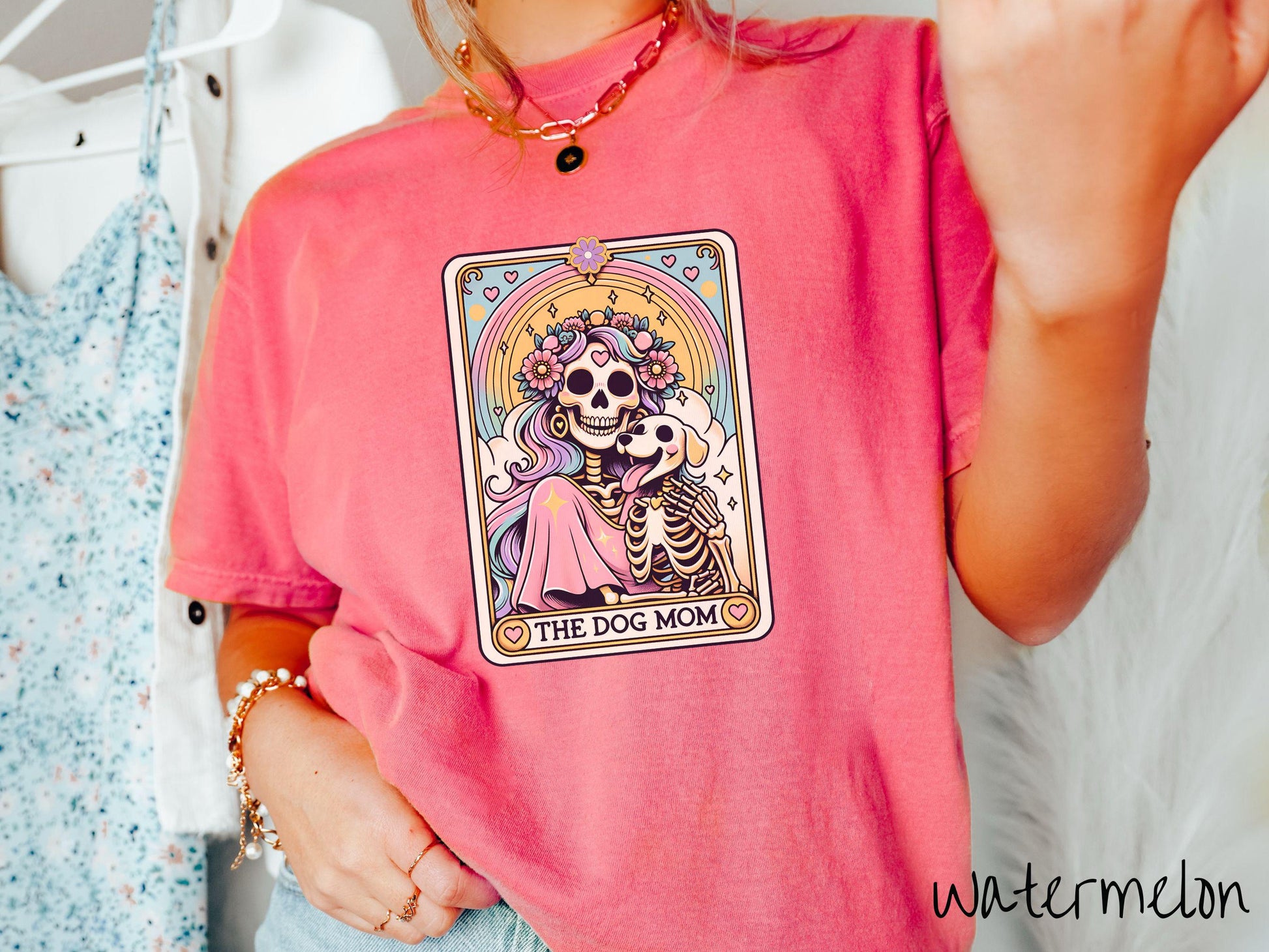 A vintage Comfort Colors t-shirt features a tarot card design with the text “The Dog Mom” along the bottom, framed by red hearts. Inside the tarot card is a female skeleton with flowing hair adorned with colorful flowers holding a skeleton dog.