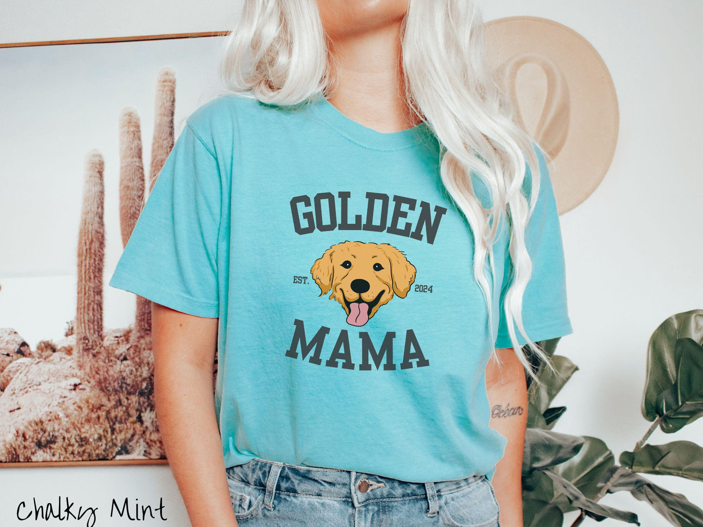 A vintage Comfort Colors t-shirt features a design with the words Golden Mama in a classic university-style font, framing an adorable black and orange Golden Retriever with its tongue out. The playful touch of EST. 2024 adds charm to the design.