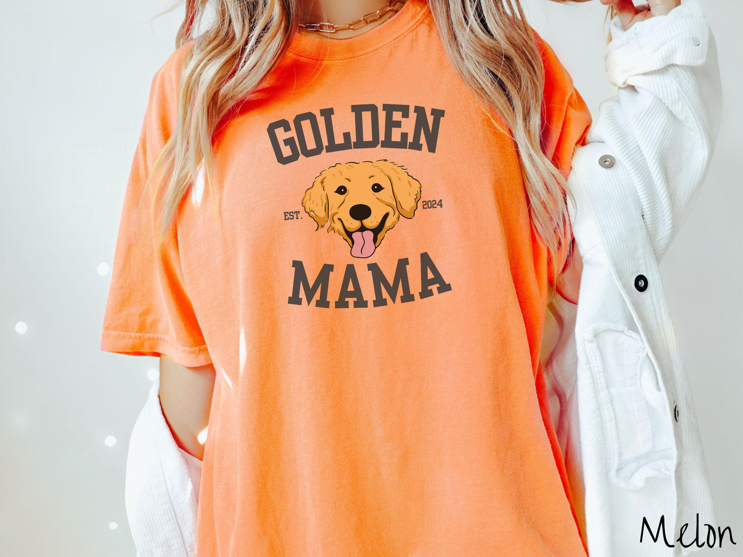 A vintage Comfort Colors t-shirt features a design with the words Golden Mama in a classic university-style font, framing an adorable black and orange Golden Retriever with its tongue out. The playful touch of EST. 2024 adds charm to the design.