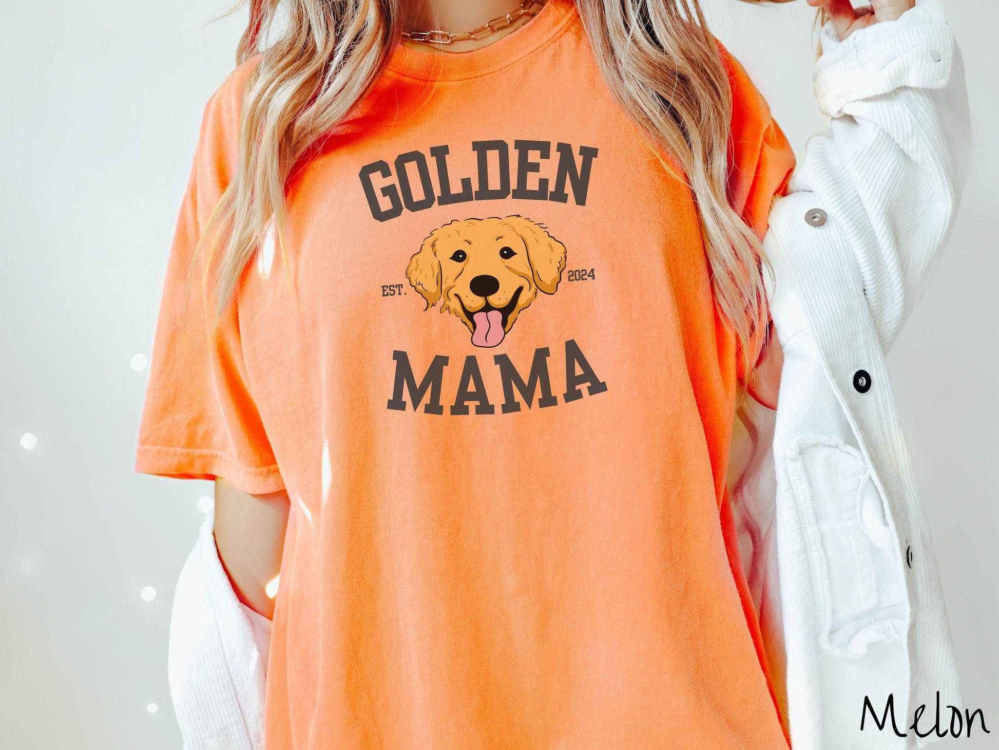 A vintage Comfort Colors t-shirt features a design with the words Golden Mama in a classic university-style font, framing an adorable black and orange Golden Retriever with its tongue out. The playful touch of EST. 2024 adds charm to the design.