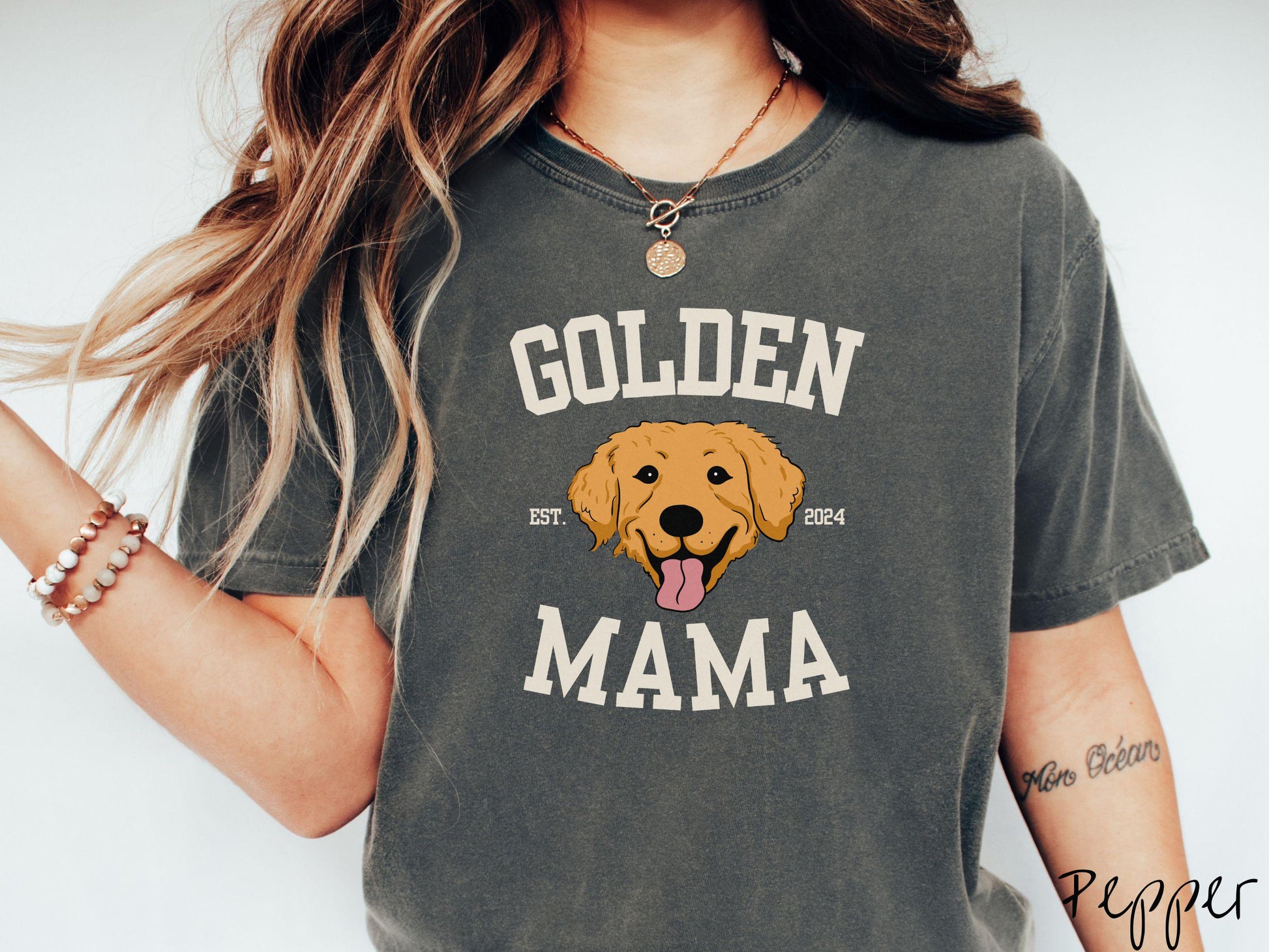 A vintage Comfort Colors t-shirt features a design with the words Golden Mama in a classic university-style font, framing an adorable black and orange Golden Retriever with its tongue out. The playful touch of EST. 2024 adds charm to the design.