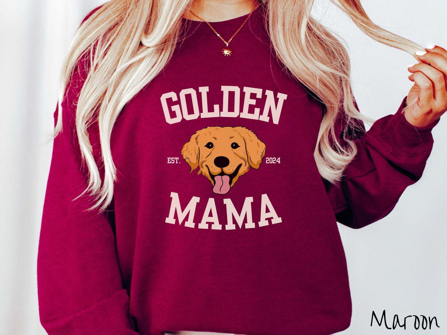 A vintage Gildan sweatshirt features a design with the words Golden Mama in a classic university-style font, framing an adorable black and orange Golden Retriever with its tongue out. The playful touch of EST. 2024 adds charm to the design.