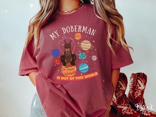 A vintage Comfort Colors t-shirt featuring the text My Doberman is Out of This World. The design showcases a Doberman sitting proudly atop Jupiter, surrounded by a vibrant solar system filled with colorful planets and twinkling stars.