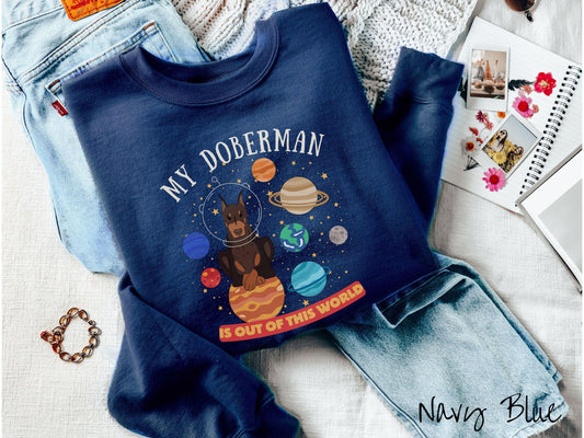 A vintage Gildan sweatshirt featuring the text My Doberman is Out of This World. The design showcases a Doberman sitting proudly atop Jupiter, surrounded by a vibrant solar system filled with colorful planets and twinkling stars.