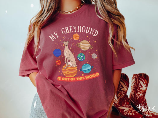 A vintage Comfort Colors t-shirt featuring the text My Greyhound is Out of This World. The design showcases a Greyhound sitting proudly atop Jupiter, surrounded by a vibrant solar system filled with colorful planets and twinkling stars.
