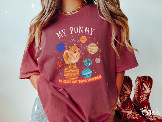 A vintage Comfort Colors t-shirt featuring the text My Pomeranian is Out of This World. The design showcases an adorable Pomeranian sitting proudly atop Jupiter, surrounded by a vibrant solar system filled with colorful planets and twinkling stars.