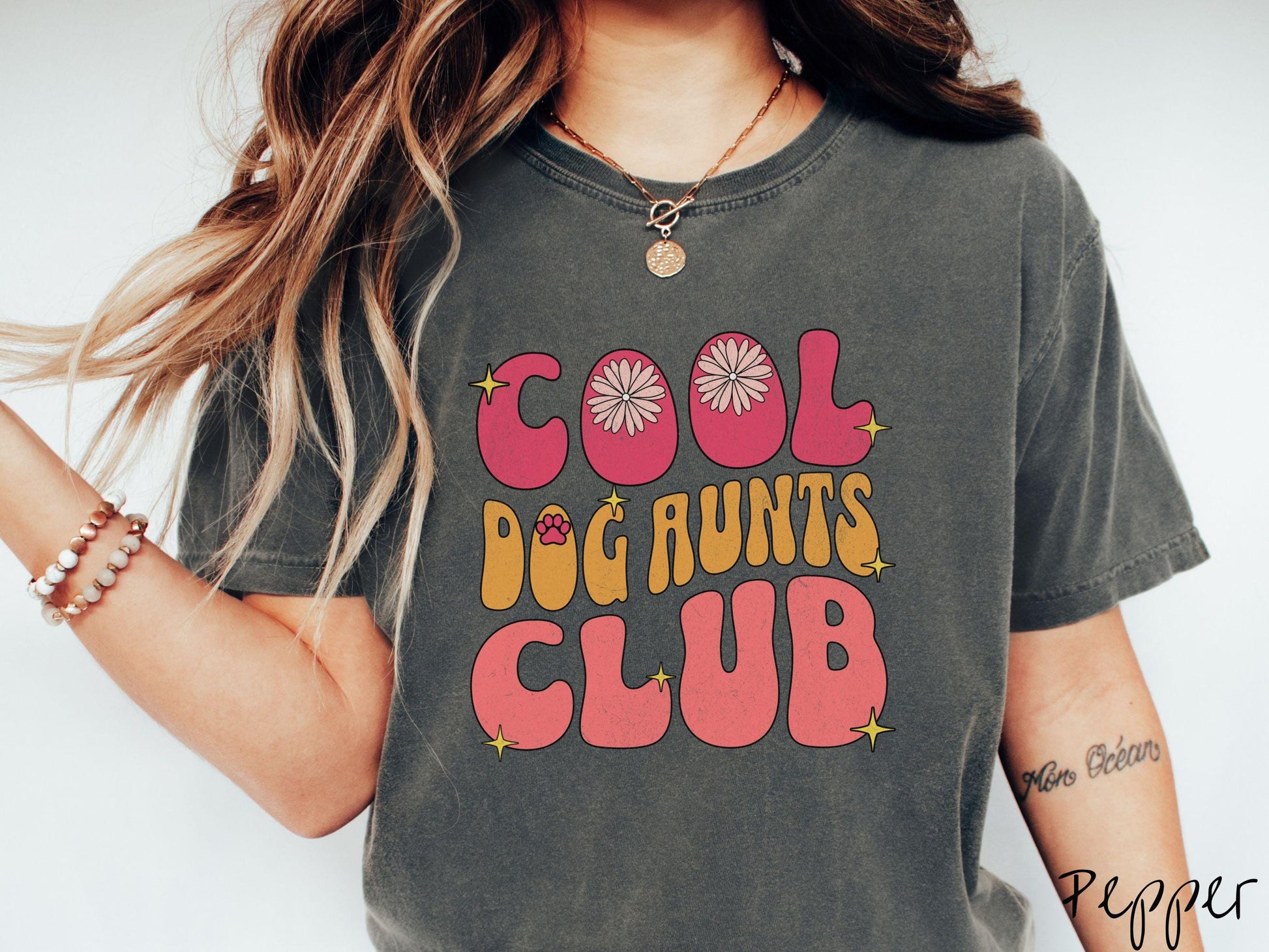 A vintage Comfort Colors t-shirt featuring the text "Cool Dog Aunts Club" in bold red, yellow, and orange fonts with flower petals nestled in the "O" of COOL, a cute red paw print in the "O" of DOG, and sparkling yellow stars throughout.