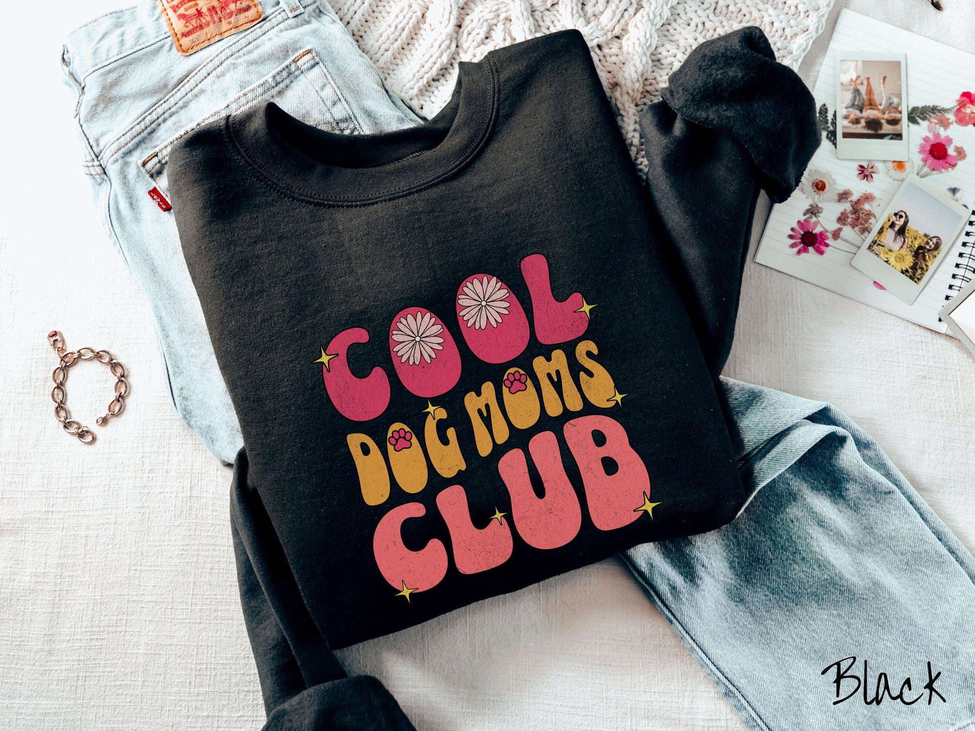 A vintage Gildan sweatshirt featuring the playful text Cool Dog Moms Club, this design is full of charming details. From flower petals nestled in the &quot;O&quot; of COOL to a cute red paw print in the &quot;O&quot; of DOG and sparkling yellow stars throughout.