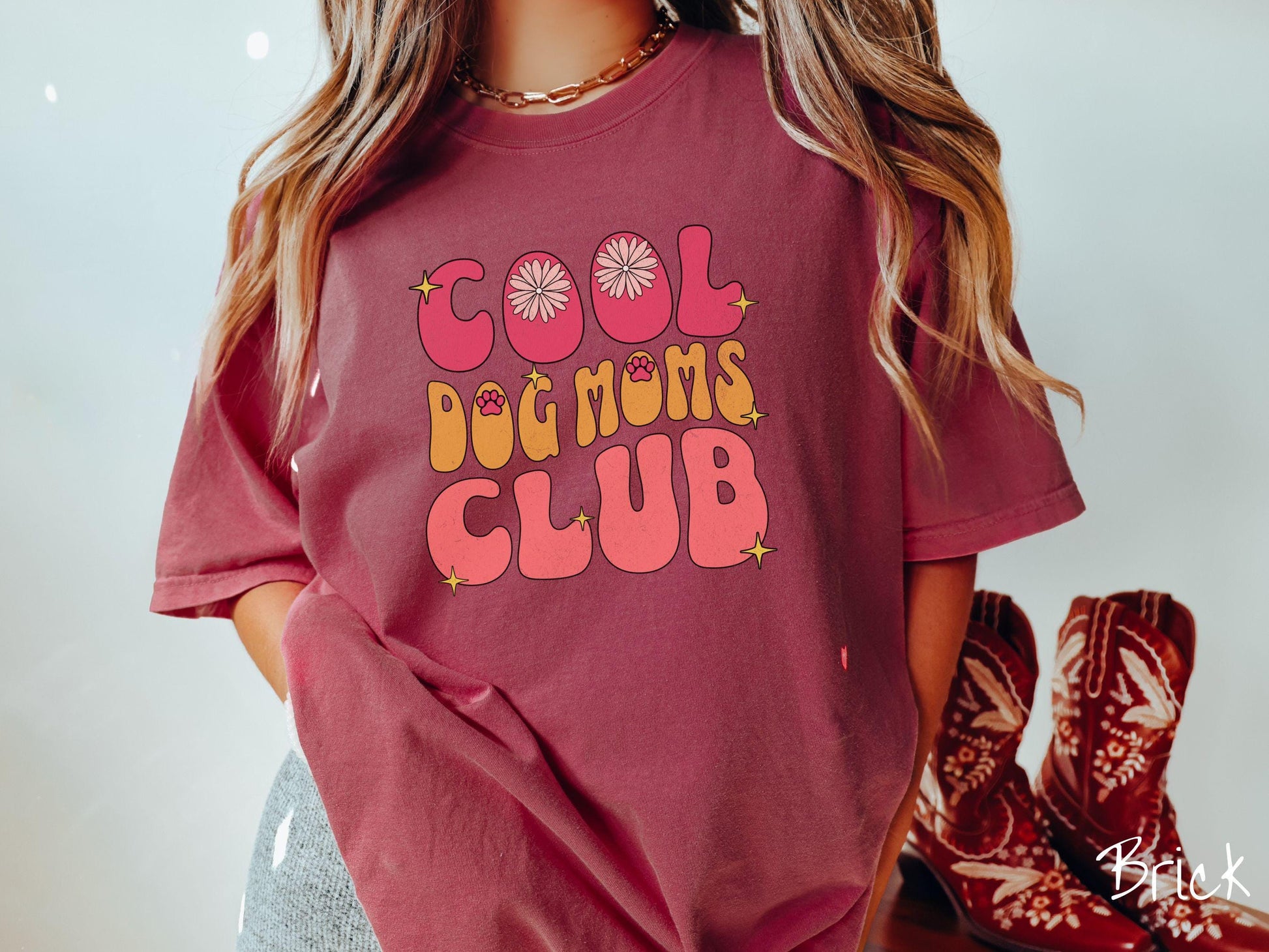 A vintage Comfort Colors t-shirt featuring the playful text Cool Dog Moms Club, this design is full of charming details. From flower petals nestled in the &quot;O&quot; of COOL to a cute red paw print in the &quot;O&quot; of DOG and sparkling yellow stars throughout.