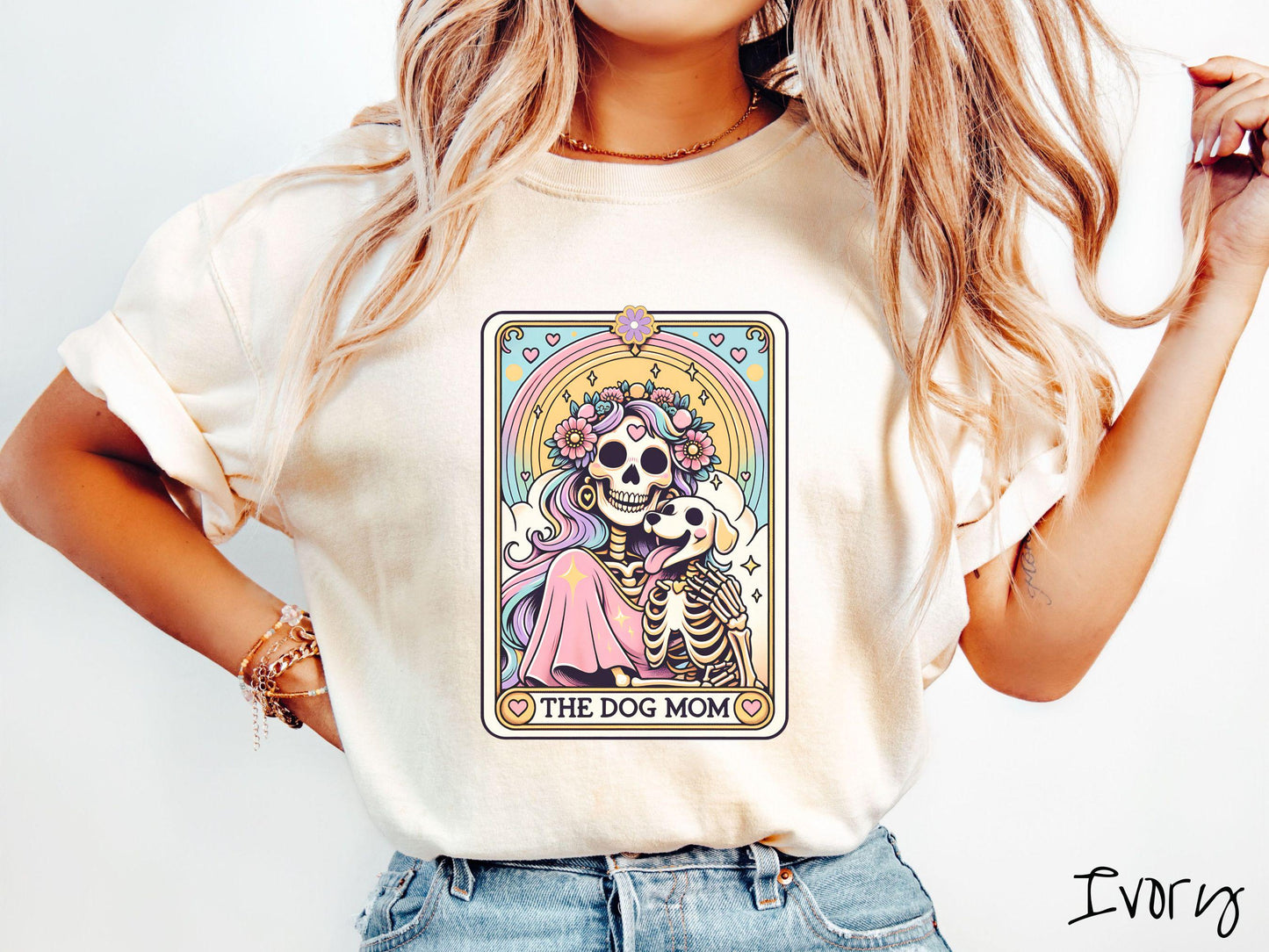 A vintage Comfort Colors t-shirt features a tarot card design with the text “The Dog Mom” along the bottom, framed by red hearts. Inside the tarot card is a female skeleton with flowing hair adorned with colorful flowers holding a skeleton dog.