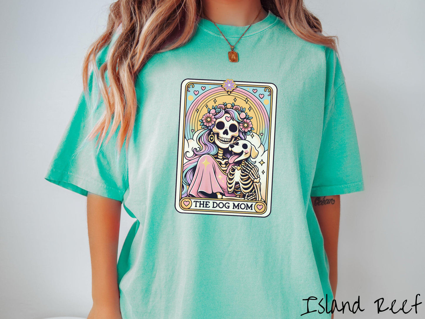 A vintage Comfort Colors t-shirt features a tarot card design with the text “The Dog Mom” along the bottom, framed by red hearts. Inside the tarot card is a female skeleton with flowing hair adorned with colorful flowers holding a skeleton dog.
