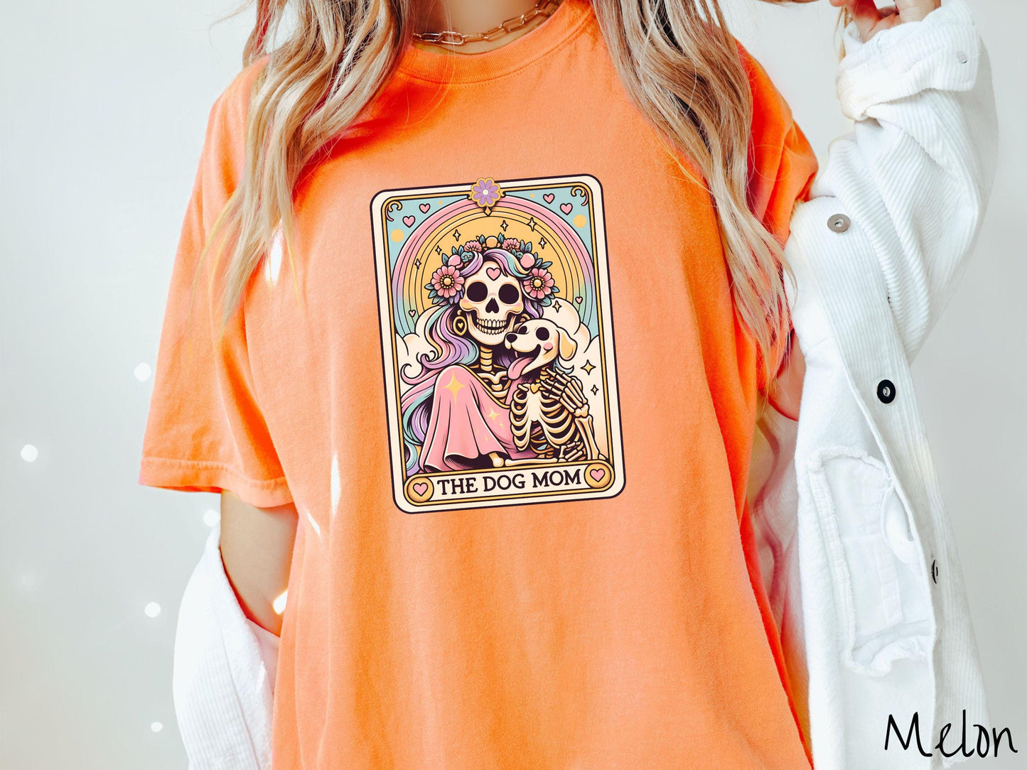 A vintage Comfort Colors t-shirt features a tarot card design with the text “The Dog Mom” along the bottom, framed by red hearts. Inside the tarot card is a female skeleton with flowing hair adorned with colorful flowers holding a skeleton dog.