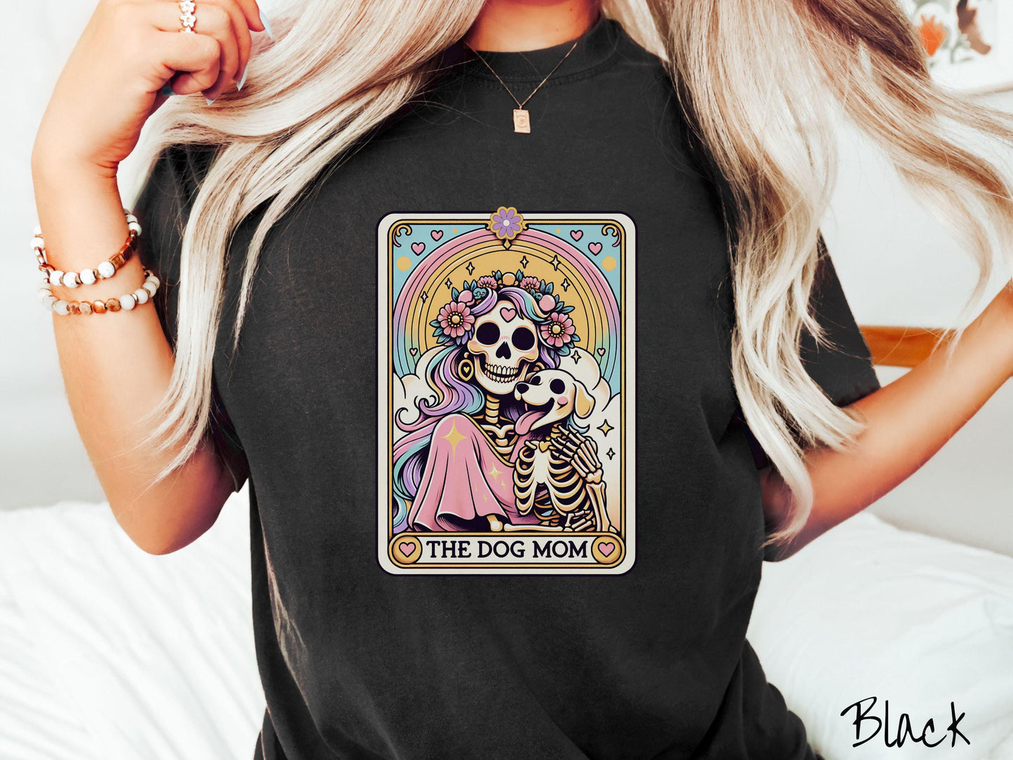 A vintage Comfort Colors t-shirt features a tarot card design with the text “The Dog Mom” along the bottom, framed by red hearts. Inside the tarot card is a female skeleton with flowing hair adorned with colorful flowers holding a skeleton dog.