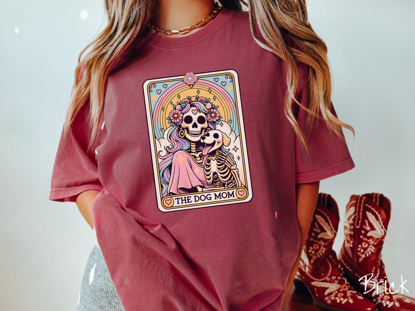A vintage Comfort Colors t-shirt features a tarot card design with the text “The Dog Mom” along the bottom, framed by red hearts. Inside the tarot card is a female skeleton with flowing hair adorned with colorful flowers holding a skeleton dog.
