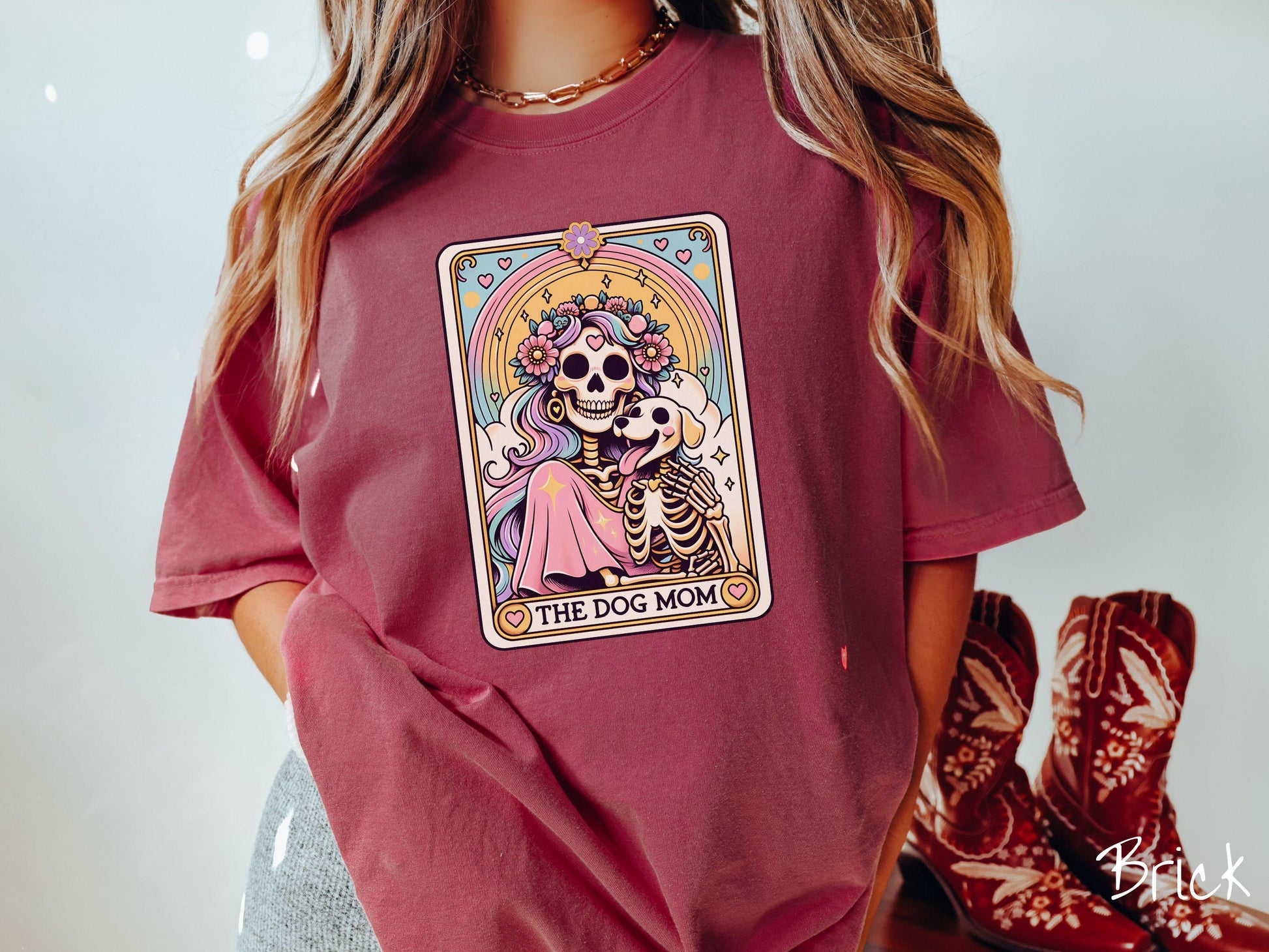 A vintage Comfort Colors t-shirt features a tarot card design with the text “The Dog Mom” along the bottom, framed by red hearts. Inside the tarot card is a female skeleton with flowing hair adorned with colorful flowers holding a skeleton dog.