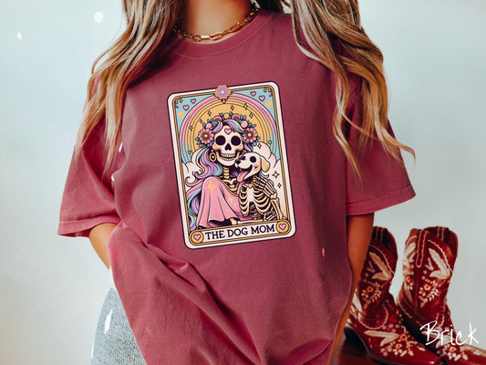A vintage Comfort Colors t-shirt features a tarot card design with the text “The Dog Mom” along the bottom, framed by red hearts. Inside the tarot card is a female skeleton with flowing hair adorned with colorful flowers holding a skeleton dog.