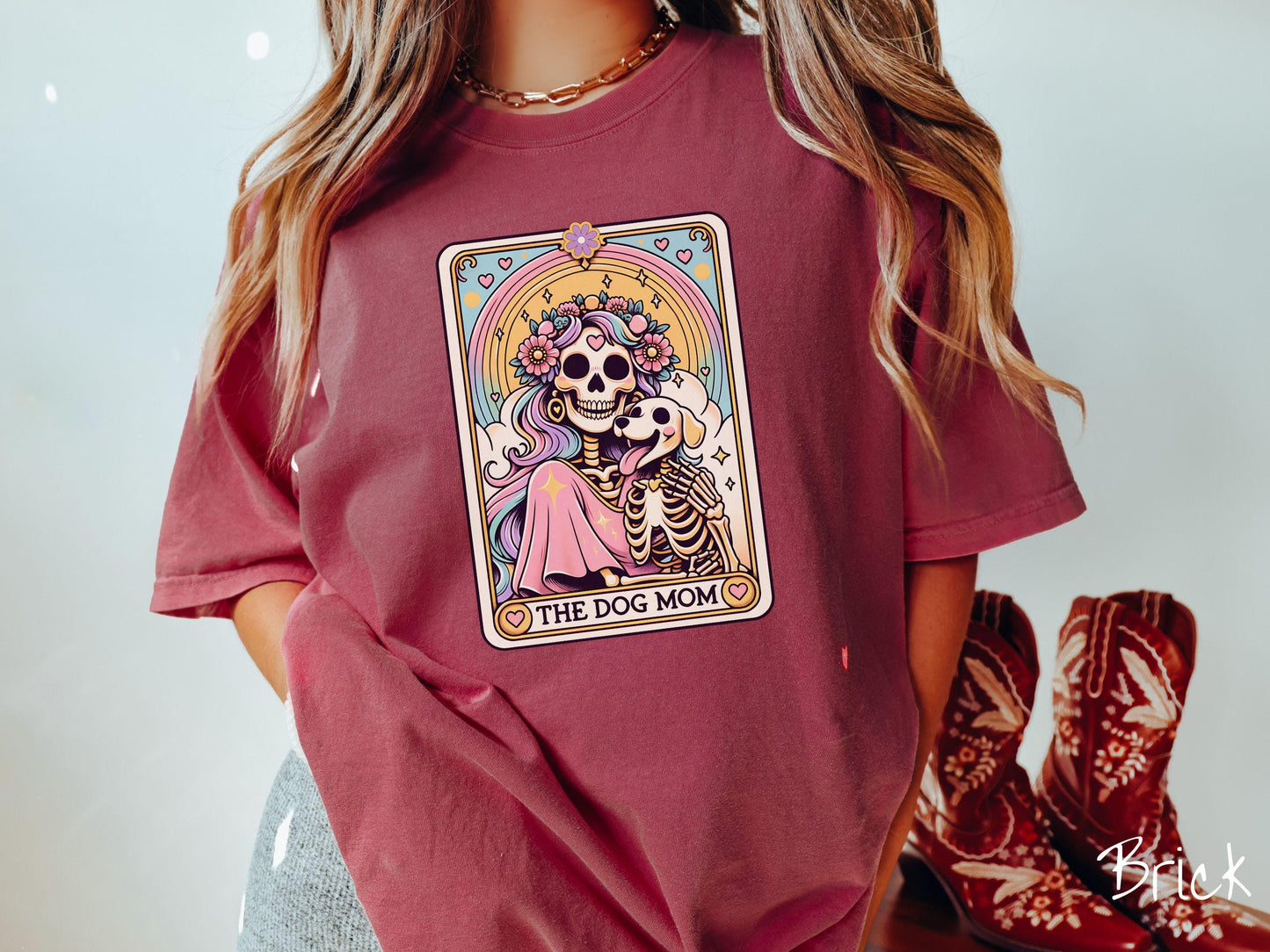 A vintage Comfort Colors t-shirt features a tarot card design with the text “The Dog Mom” along the bottom, framed by red hearts. Inside the tarot card is a female skeleton with flowing hair adorned with colorful flowers holding a skeleton dog.