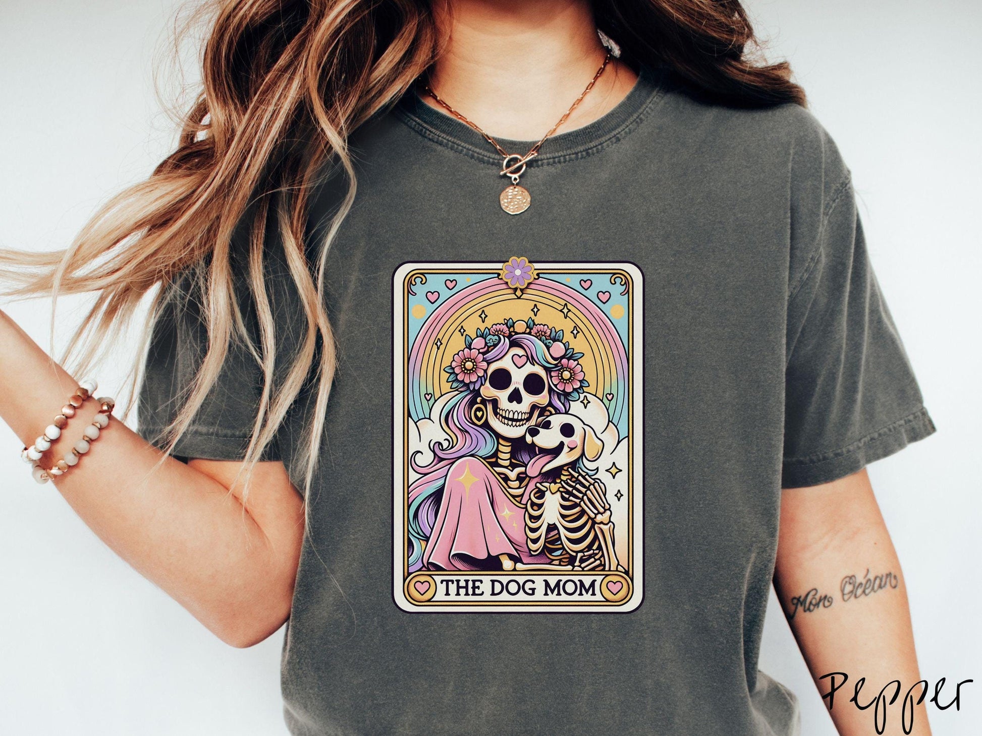 A vintage Comfort Colors t-shirt features a tarot card design with the text “The Dog Mom” along the bottom, framed by red hearts. Inside the tarot card is a female skeleton with flowing hair adorned with colorful flowers holding a skeleton dog.