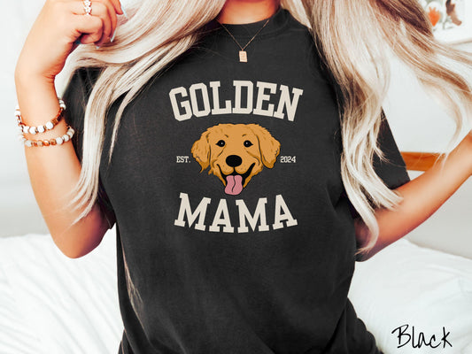 A vintage Comfort Colors t-shirt features a design with the words Golden Mama in a classic university-style font, framing an adorable black and orange Golden Retriever with its tongue out. The playful touch of EST. 2024 adds charm to the design.